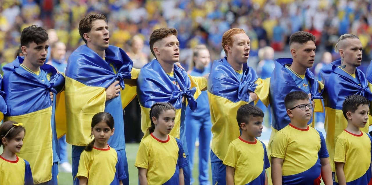 Los jugadores de la selección ucraniana con la bandera de su país atada al cuello entonan el himno nacional en su debut en la Eurocopa 2024 contra Rumanía. EFE/EPA/RONALD WITTEK